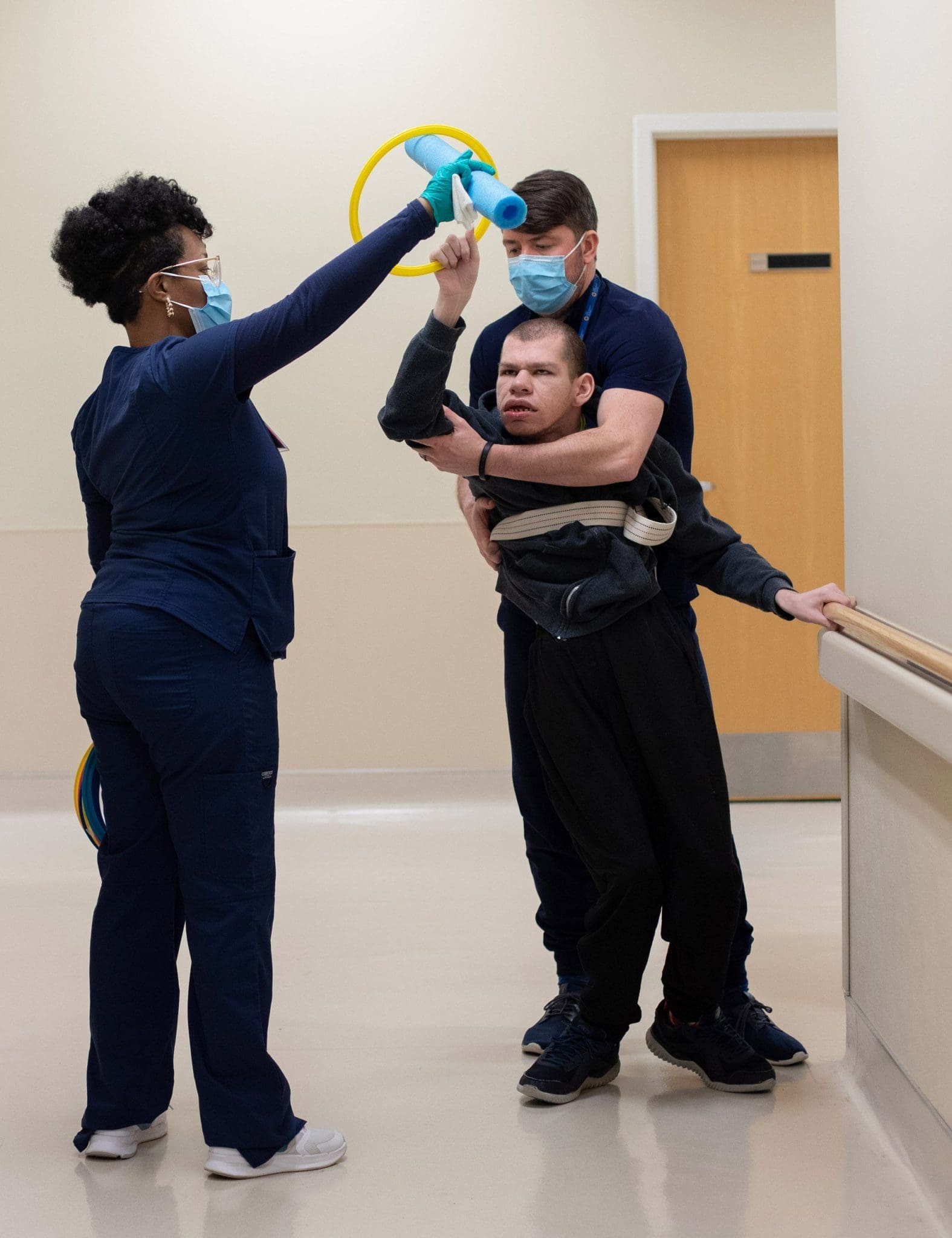 Two therapists assist a patient with intellectual and developmental disabilities (IDD) in a walking exercise down the hallway, using therapy tools to encourage mobility, coordination, and engagement.