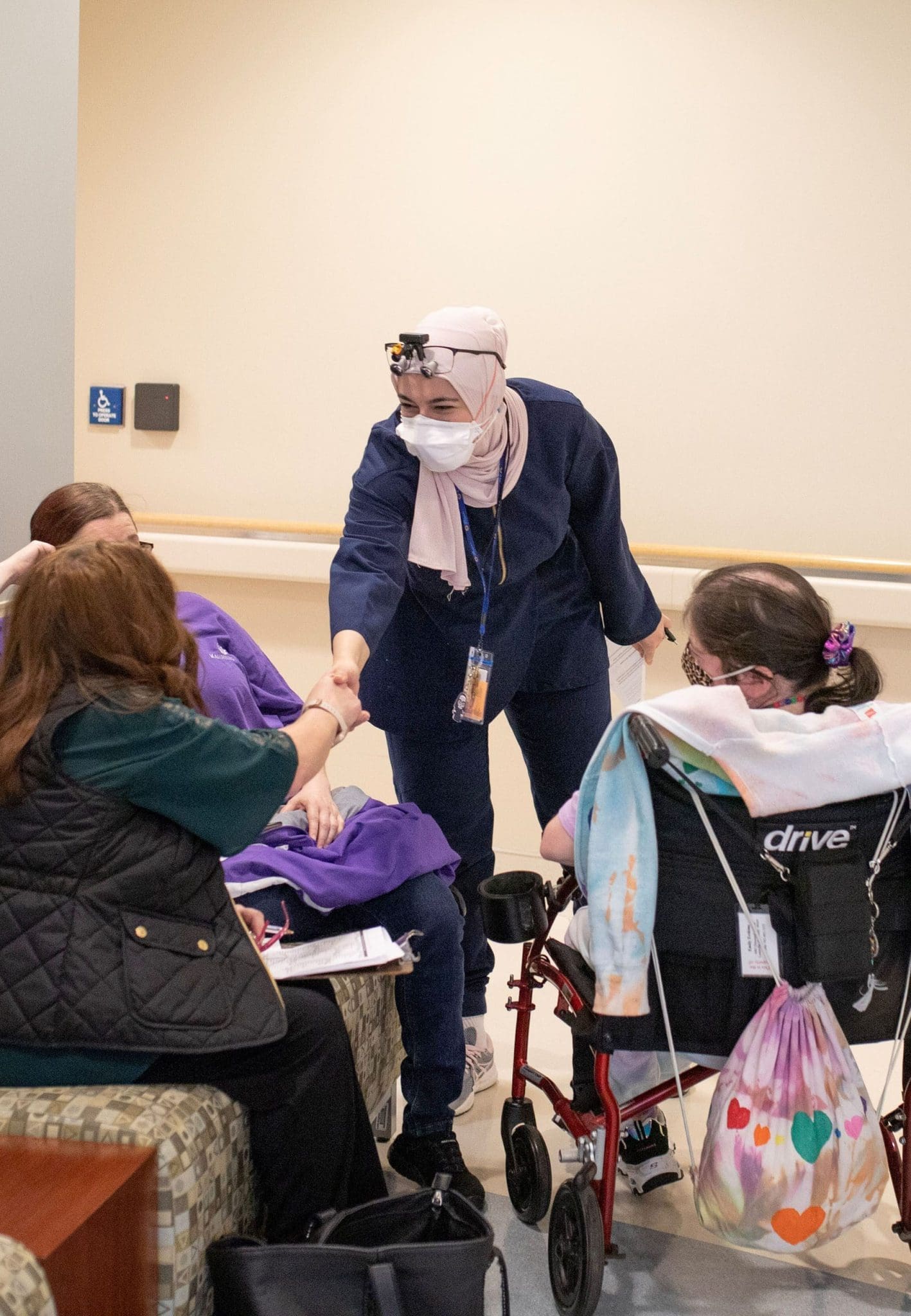 A hijab-wearing dentist extends a handshake to a seated caregiver, social worker, and a patient in a wheelchair as they await their dental appointment.