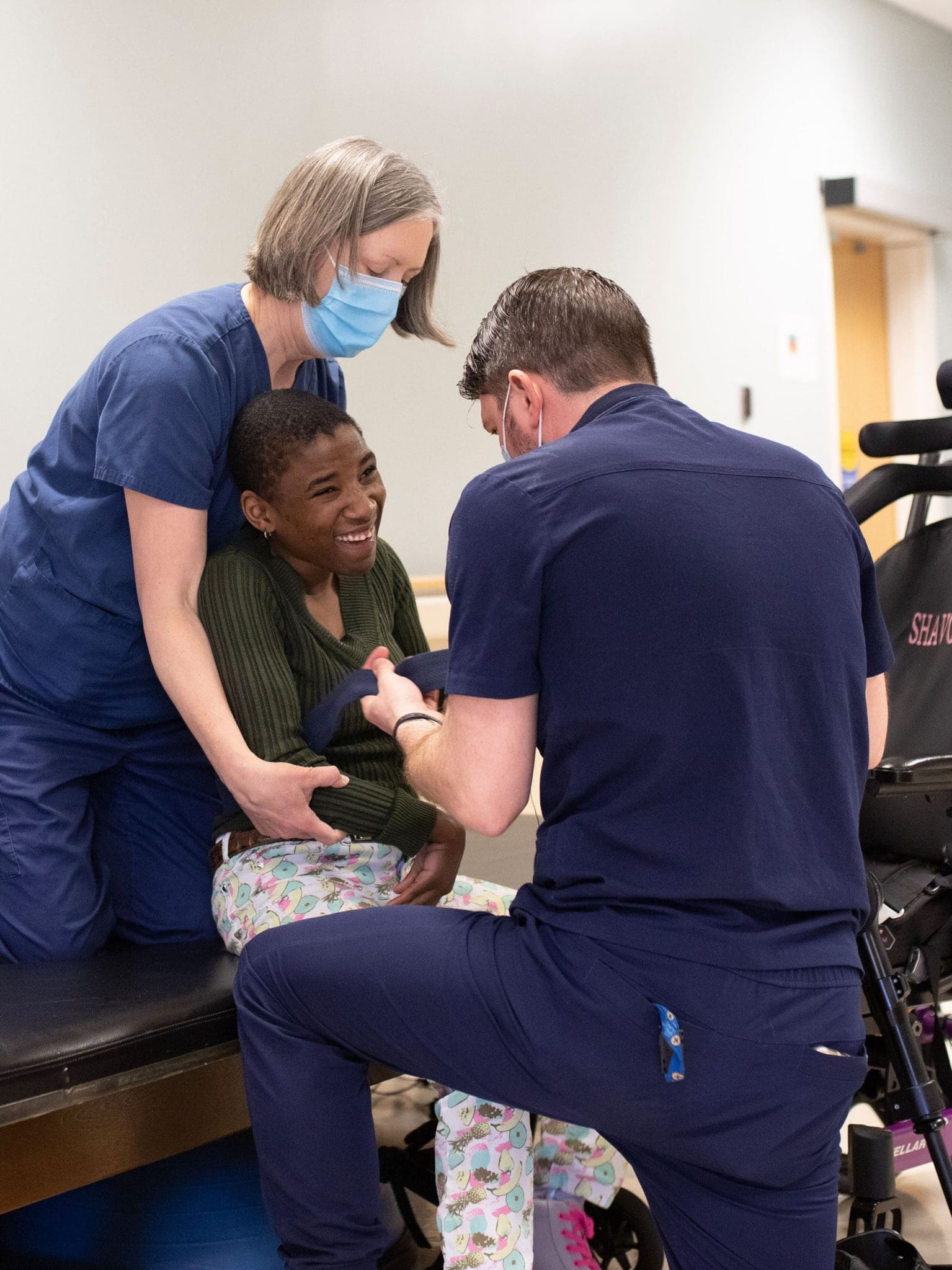 A patient with a neurodevelopmental condition smiles as two physical and occupational therapists assist her with mobility exercises, enhancing strength, independence, and daily living skills.
