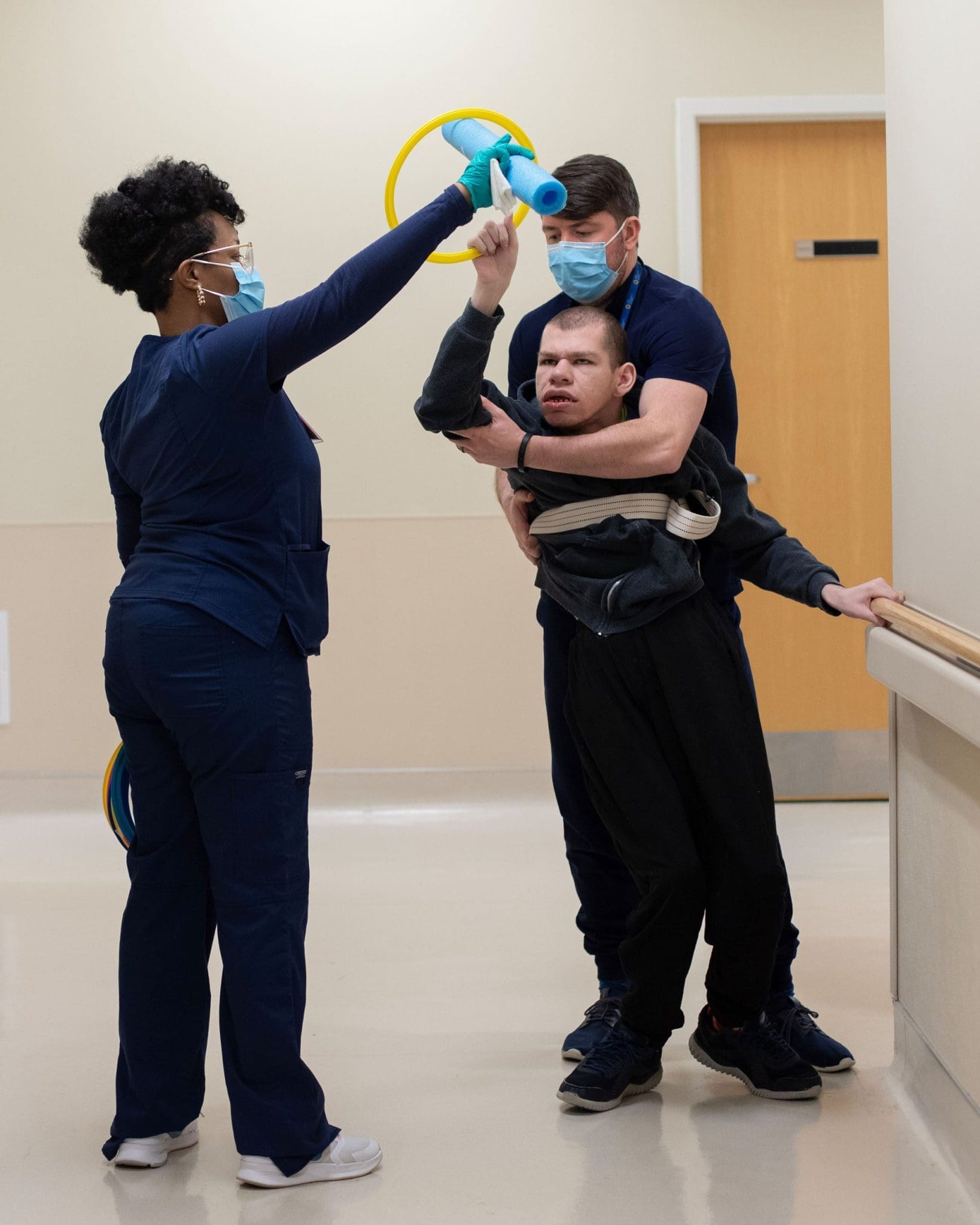 Two therapists assist a patient with intellectual and developmental disabilities (IDD) in a walking exercise down the hallway, using therapy tools to encourage mobility, coordination, and engagement.